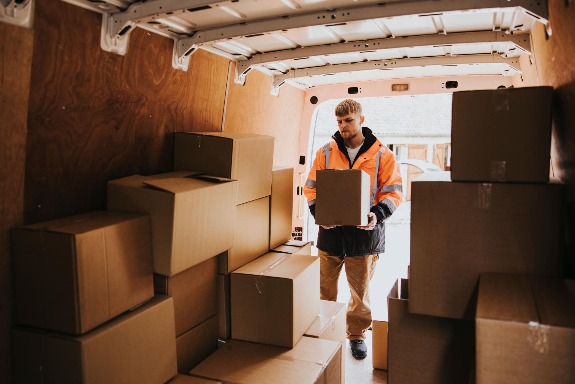 A delivery worker in a bright orange safety jacket loads a cardboard box into the back of a van.