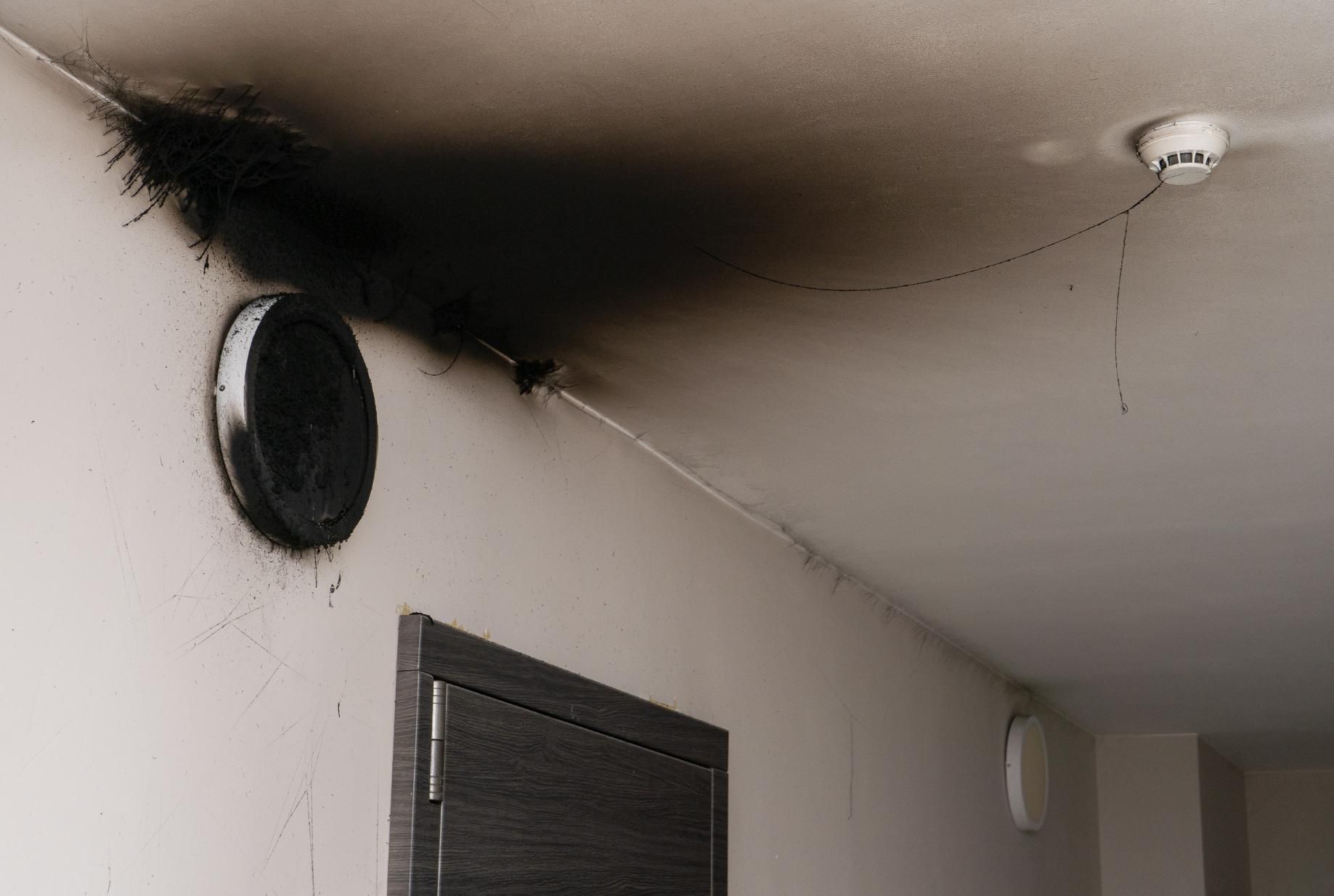 A white wall and ceiling with a large, dark soot stain radiating from a circular vent.