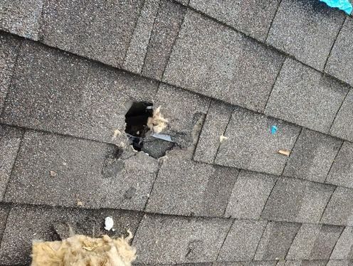 A large hole in a gray asphalt shingle roof revealing insulation and dark space underneath.