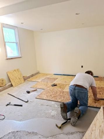 A person kneels on a concrete floor while installing plywood subflooring in a room.