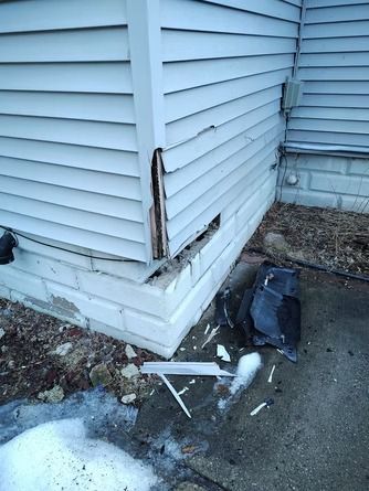 Damaged house exterior with torn grey siding, exposed wood corner, and debris.