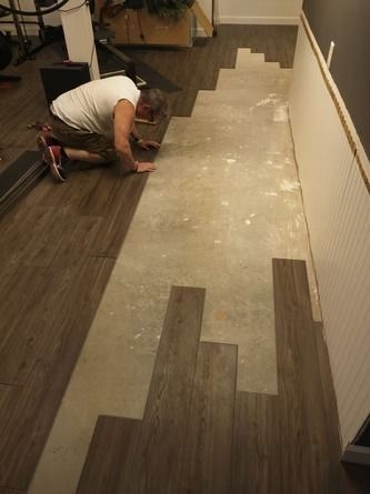A person installs brown wood-look vinyl flooring on a concrete subfloor in a room with white wainscoting.