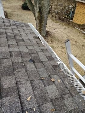 A high-angle view of a gray asphalt shingle roof with a metal gutter guard, with a ladder leaning against the eaves.