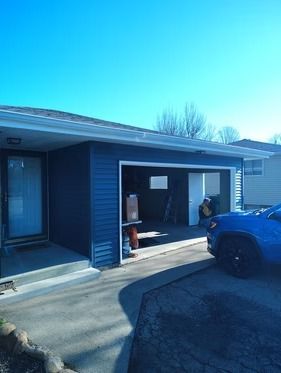 A blue-sided house with an open garage and a concrete driveway, parked next to a blue car under a clear sky.