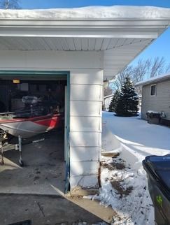 A red and white boat parked in an open garage with snow on the roof and ground outside.