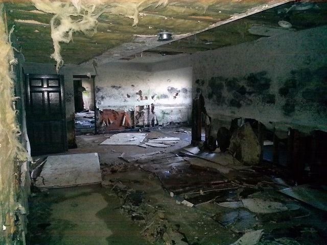 A dimly lit, dilapidated room with peeling ceiling insulation, dark debris on the floor, and an open doorway.