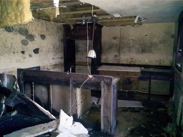 A dilapidated interior room with a wooden bar, exposed ceiling beams, insulation, and debris on walls and floor.