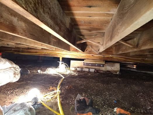 A low-angle view of a dirt crawlspace showing wooden floor joists, a support beam on a concrete pier, and wiring.