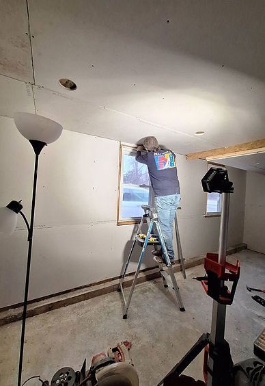 A person wearing a grey shirt works on the frame of a window in a room under construction.