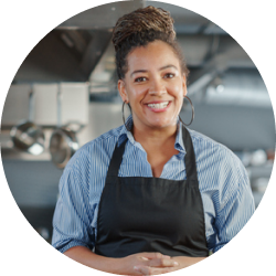 A woman in an apron is smiling in a kitchen.
