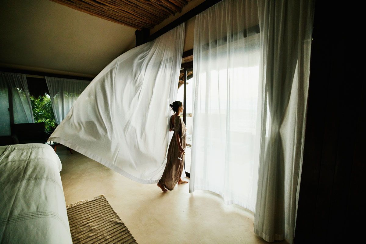 A woman in a long dress is opening a window with white curtains.