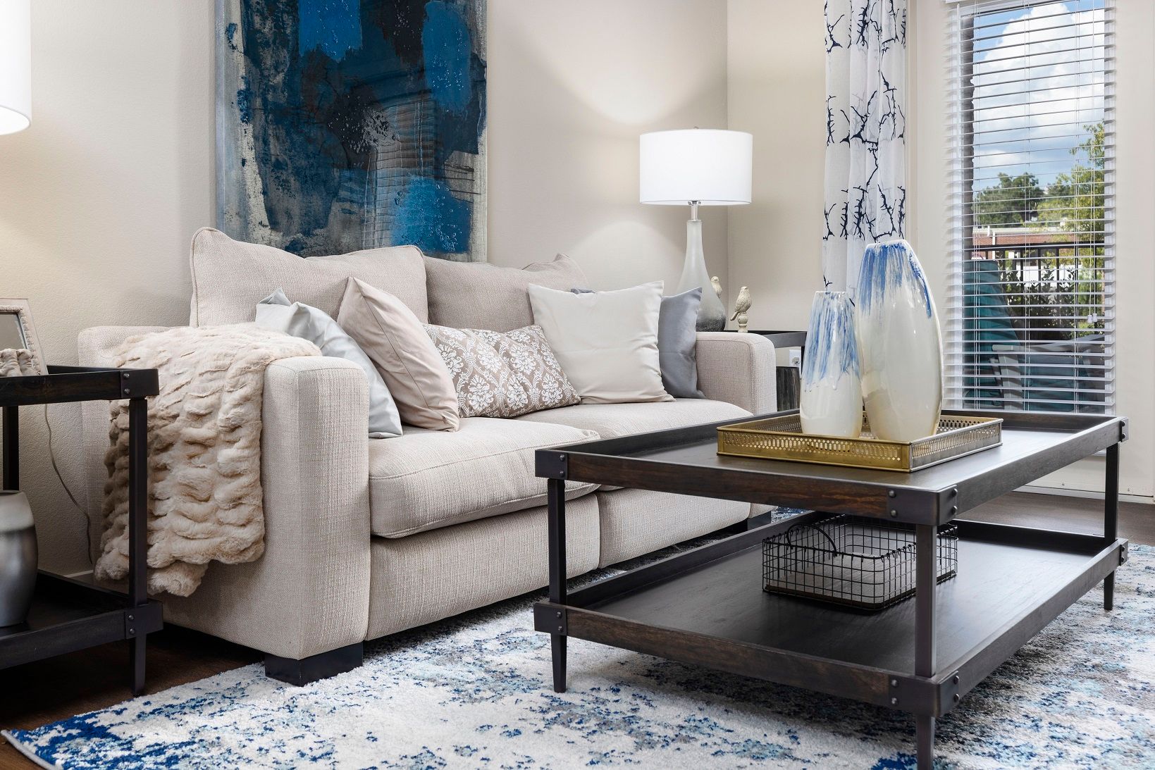 Beige sofa with blue accents in a living room, coffee table, rug, art, and window.