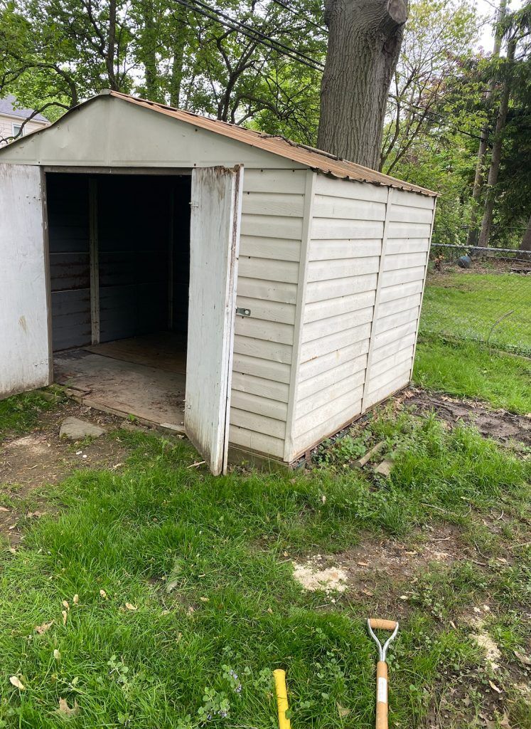 White shed with open door on green grass, shovel in the foreground.