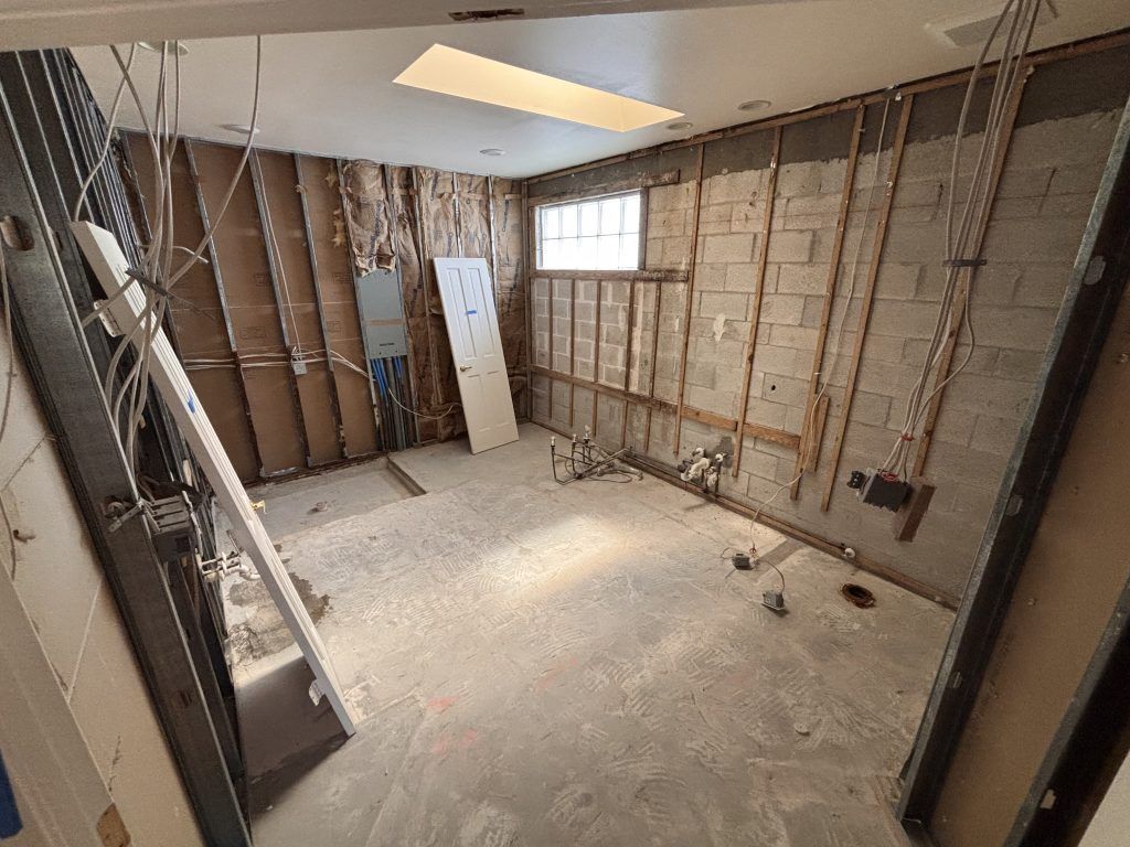 Room under construction, exposed walls and utilities. Light-colored concrete floor. Doorway on the right.