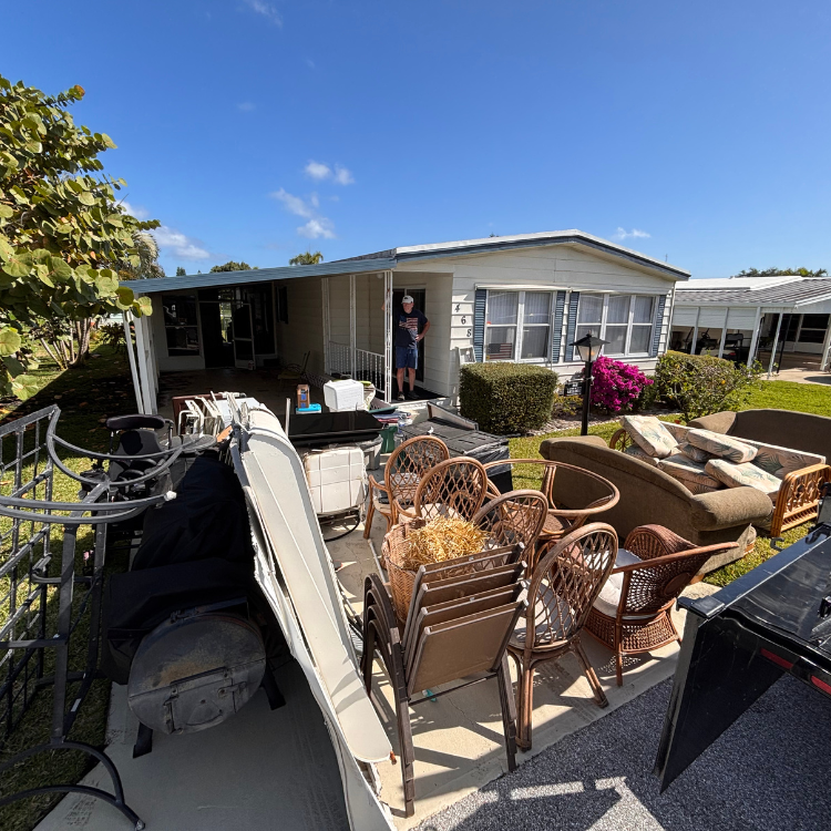 A single-story house with a man standing in the doorway is surrounded by various pieces of outdoor furniture on a sunny day.