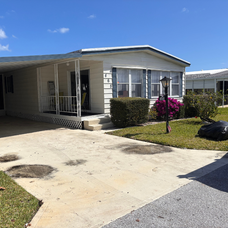 Mobile home with carport, white siding, and blue shutters, with small front yard and concrete driveway.