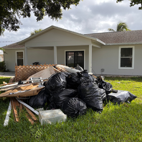 A pile of black trash bags and debris sits in front of a light gray house.