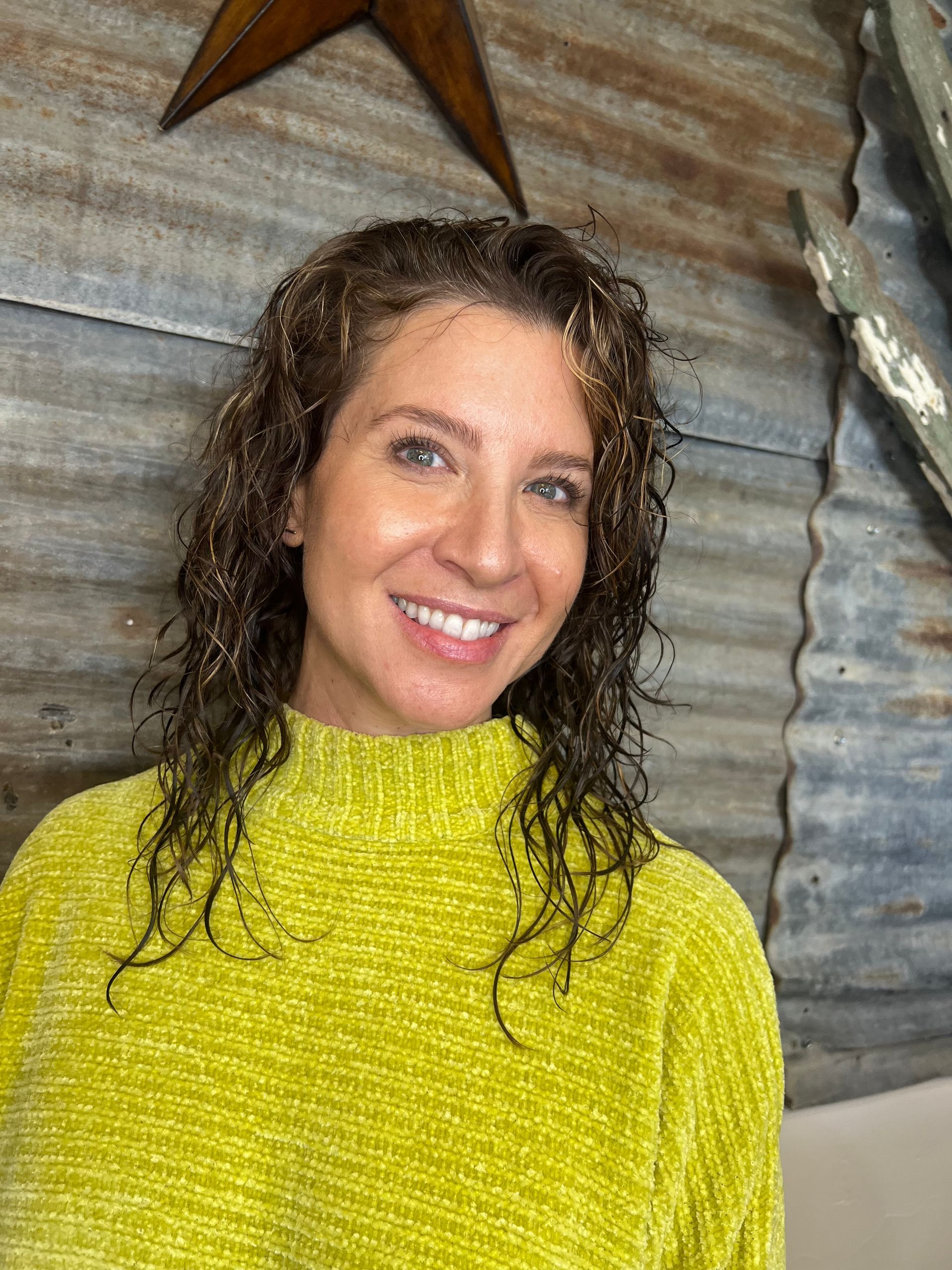A woman with curly hair is wearing a yellow sweater and smiling.