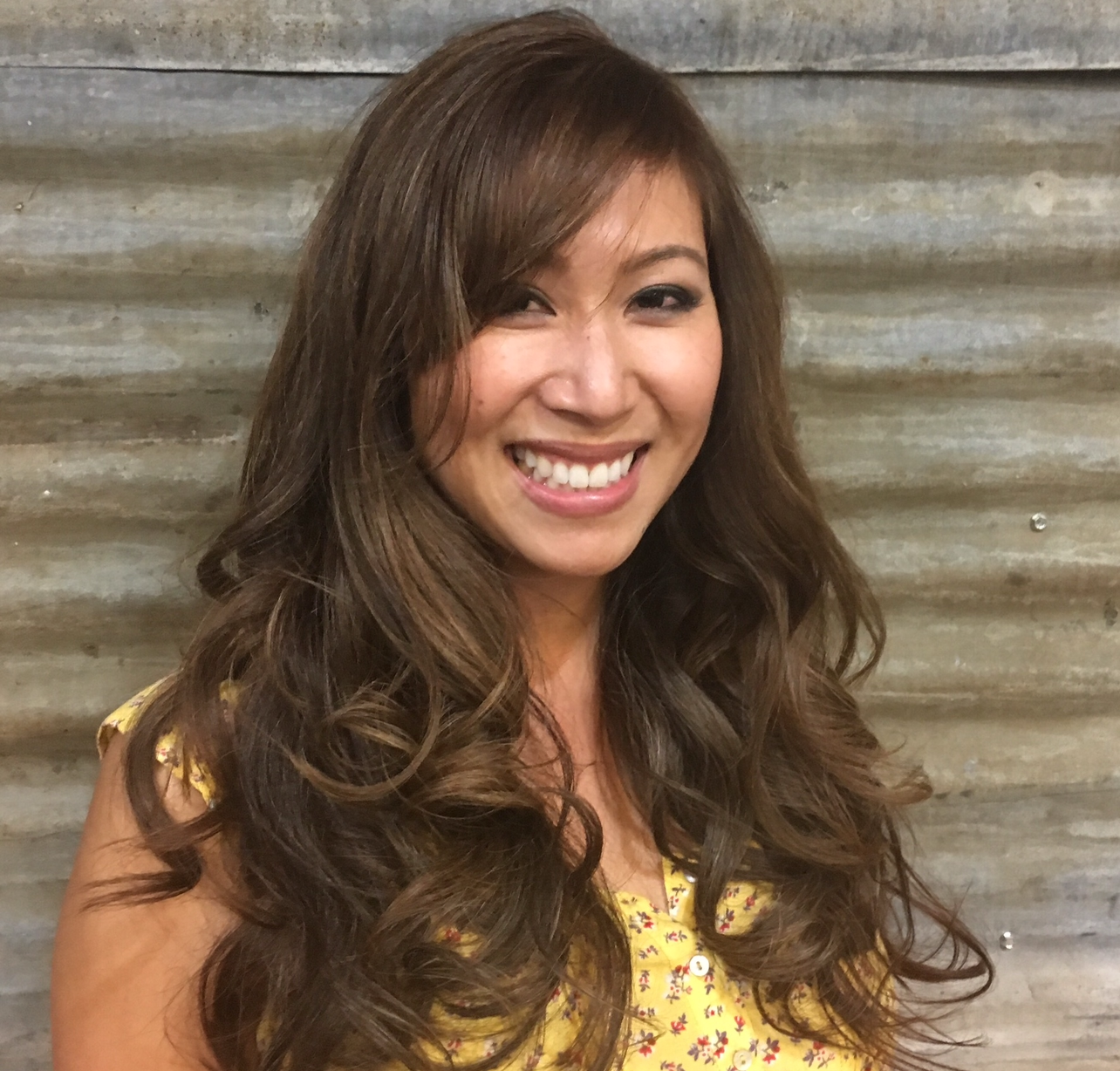 A woman with long brown hair is smiling and wearing a yellow shirt.