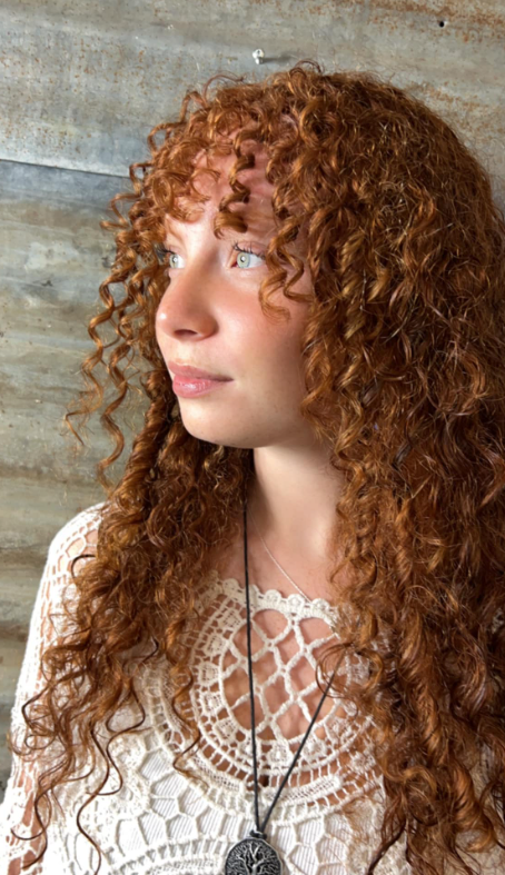 A woman with red curly hair is wearing a white lace top and a necklace.
