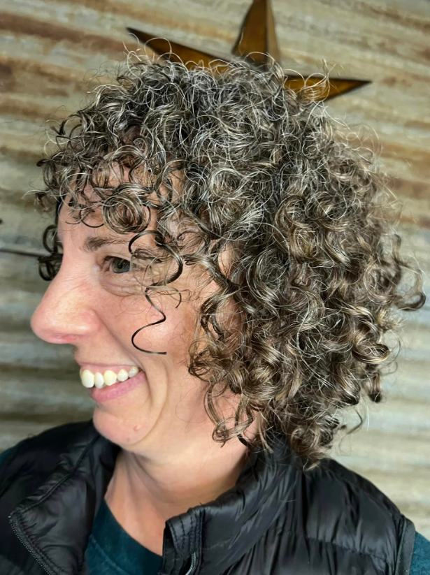 A woman with curly hair is smiling and wearing a black vest.
