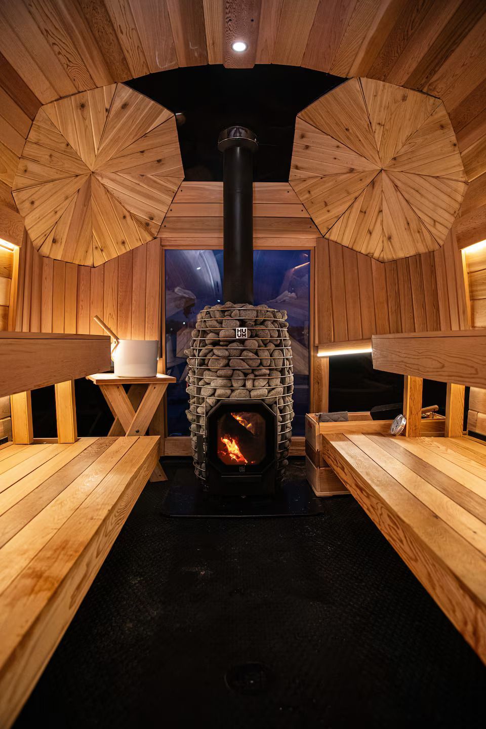 Wood-paneled sauna with wooden benches, a central stone stove, and two umbrella-like wall decorations