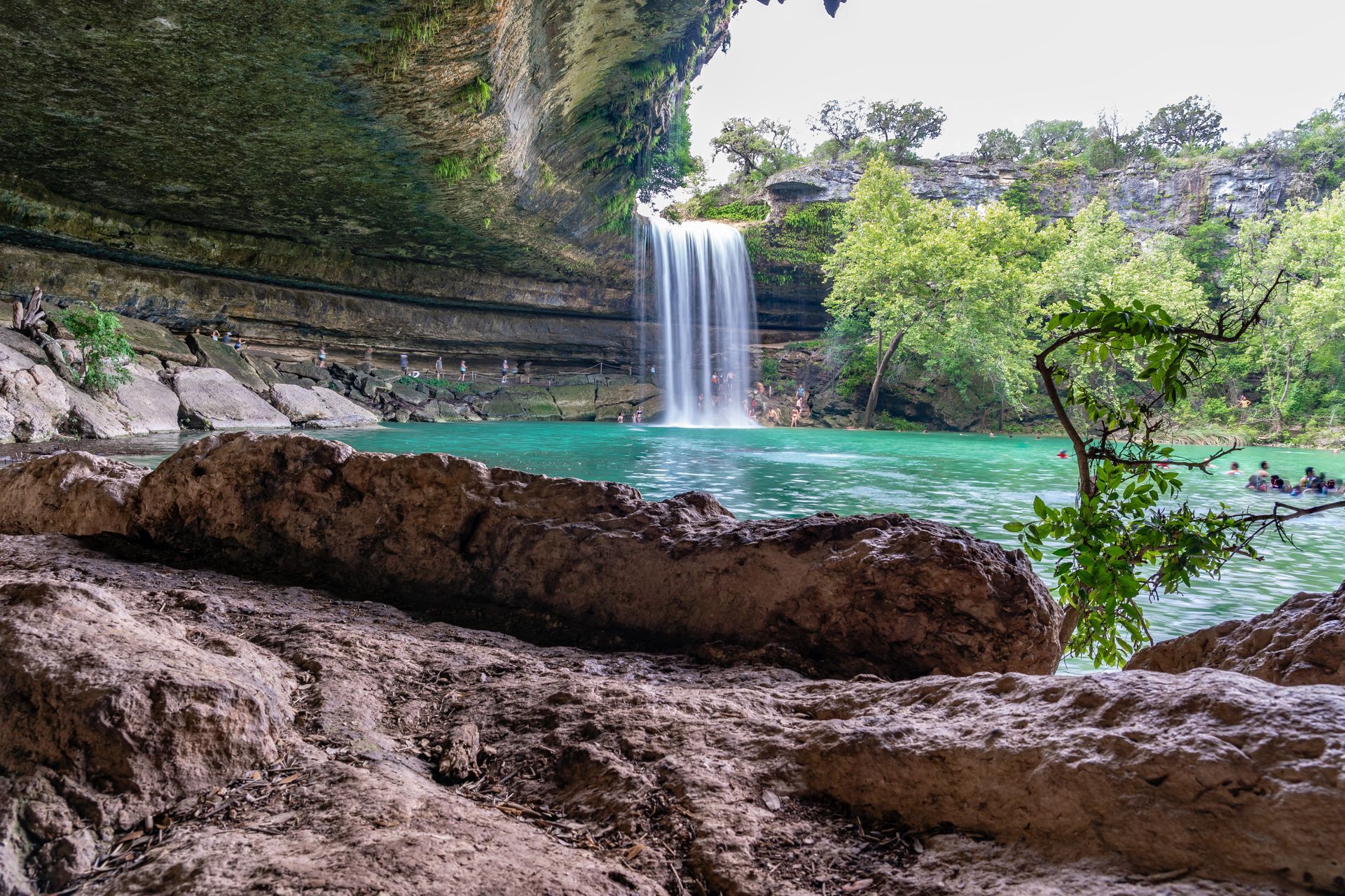 Waterfall cascading into a turquoise pool beneath a rocky overhang, with lush green trees surrounding it