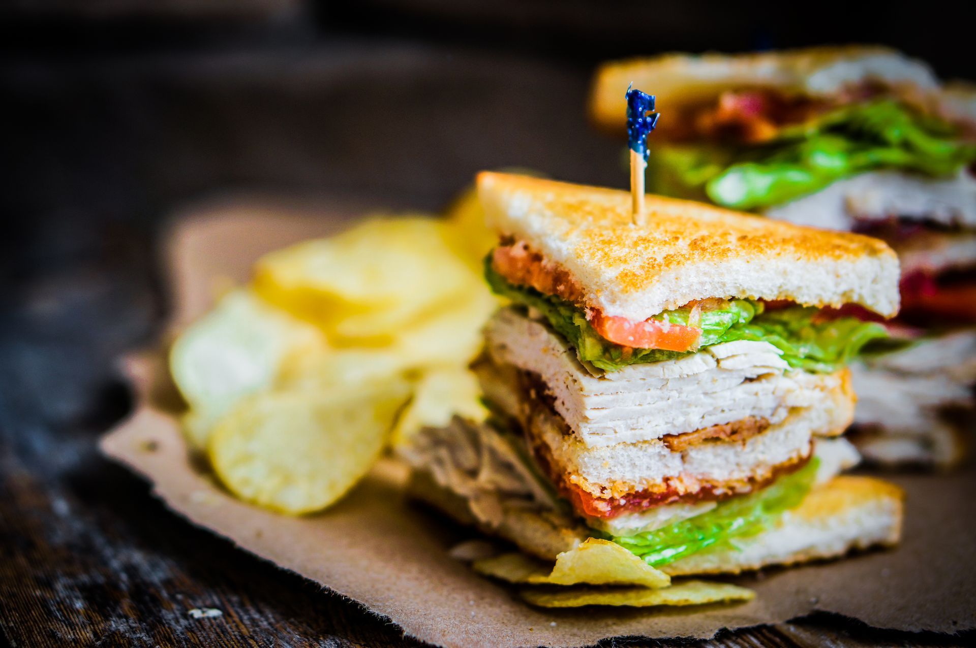 Stacked sandwich with chips on a dark plate, topped with a toothpick.