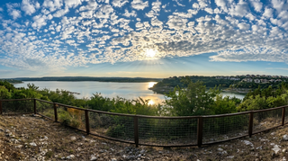 Scenic lake at sunset under a sky of scattered clouds, viewed from a hillside overlook with a fence in the foreground