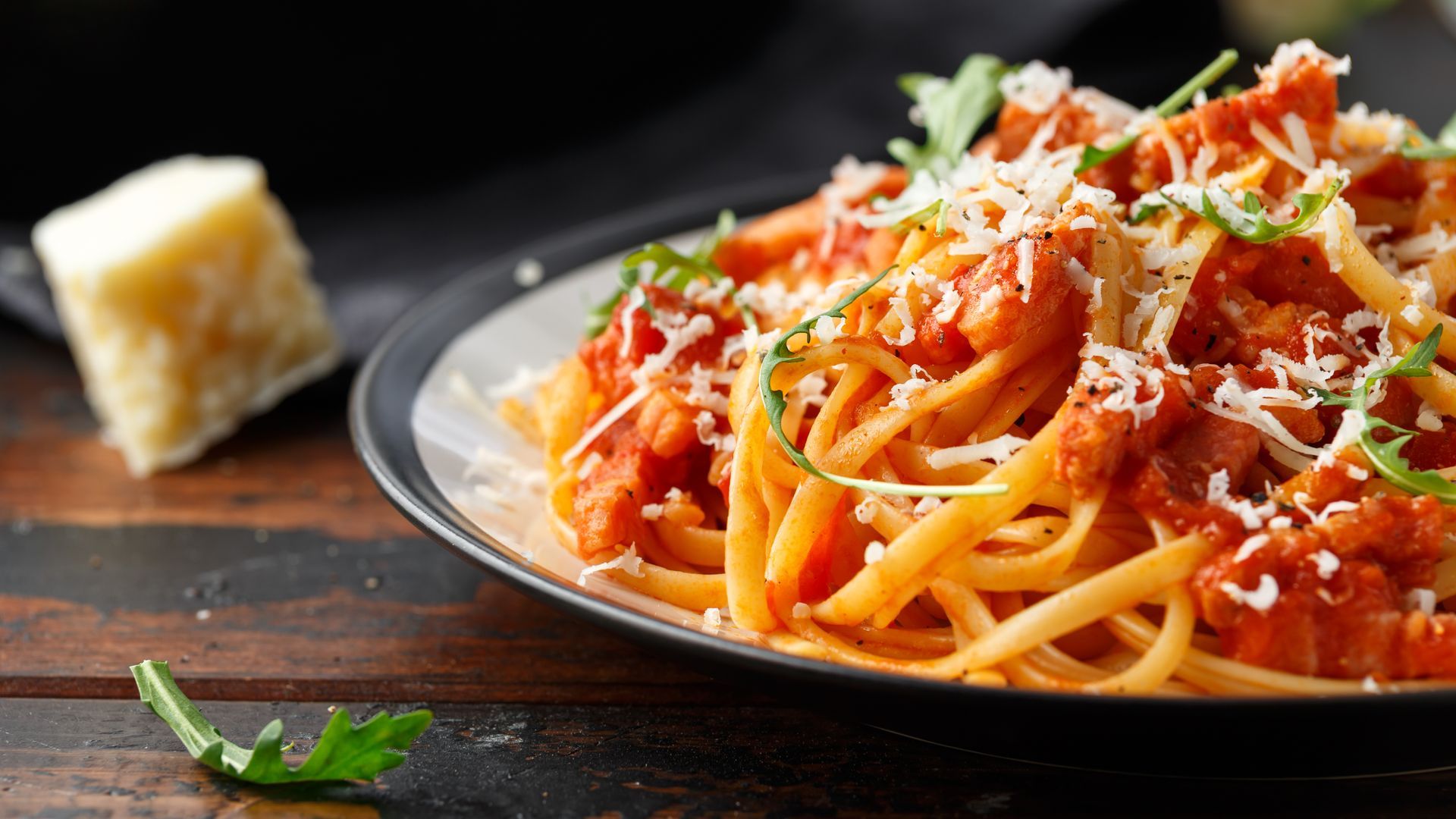 Plate of spaghetti with tomato sauce and grated cheese, with a Parmesan wedge on a dark wooden table.