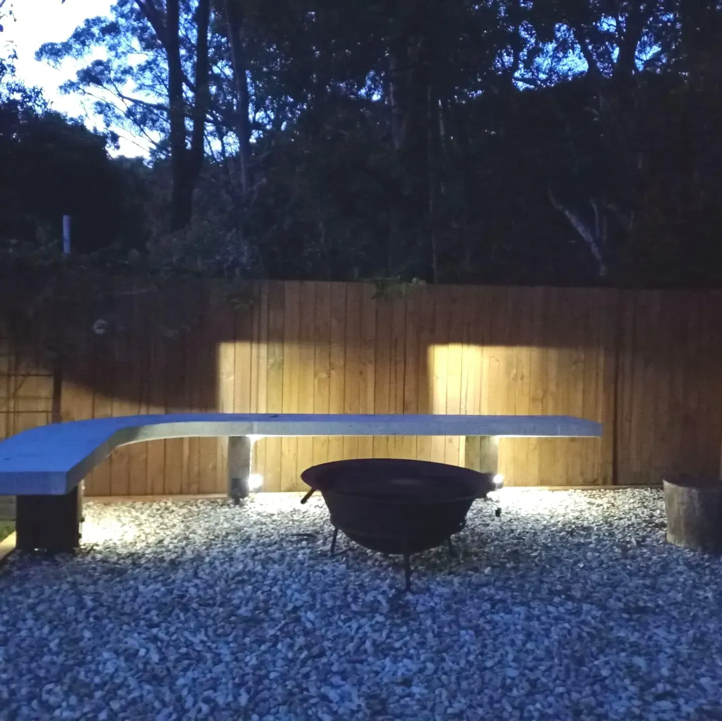 A Wooden Fence Behind a Gravel Area With a Bench and a Fire Pit — Bangalow Electrical in Bangalow, NSW