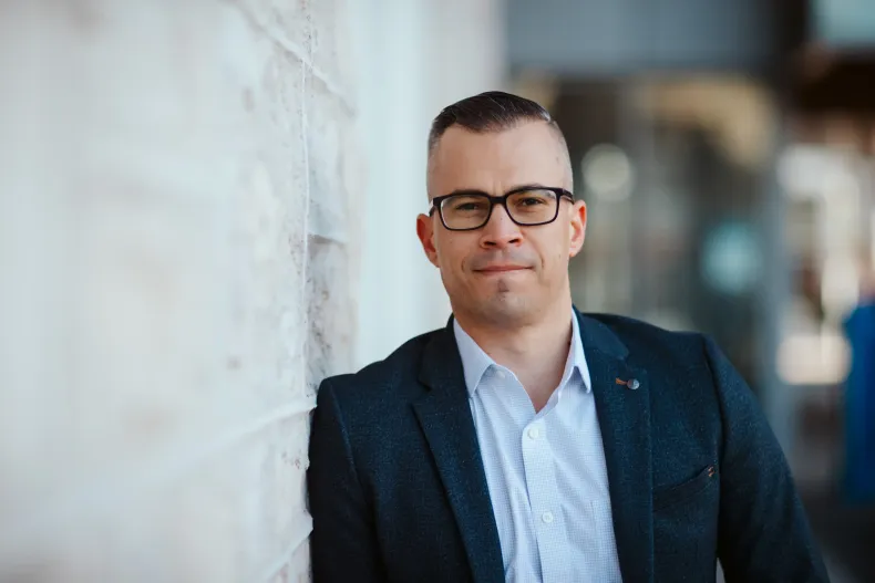 Man in glasses and a blazer leaning against a white wall, looking at the camera.