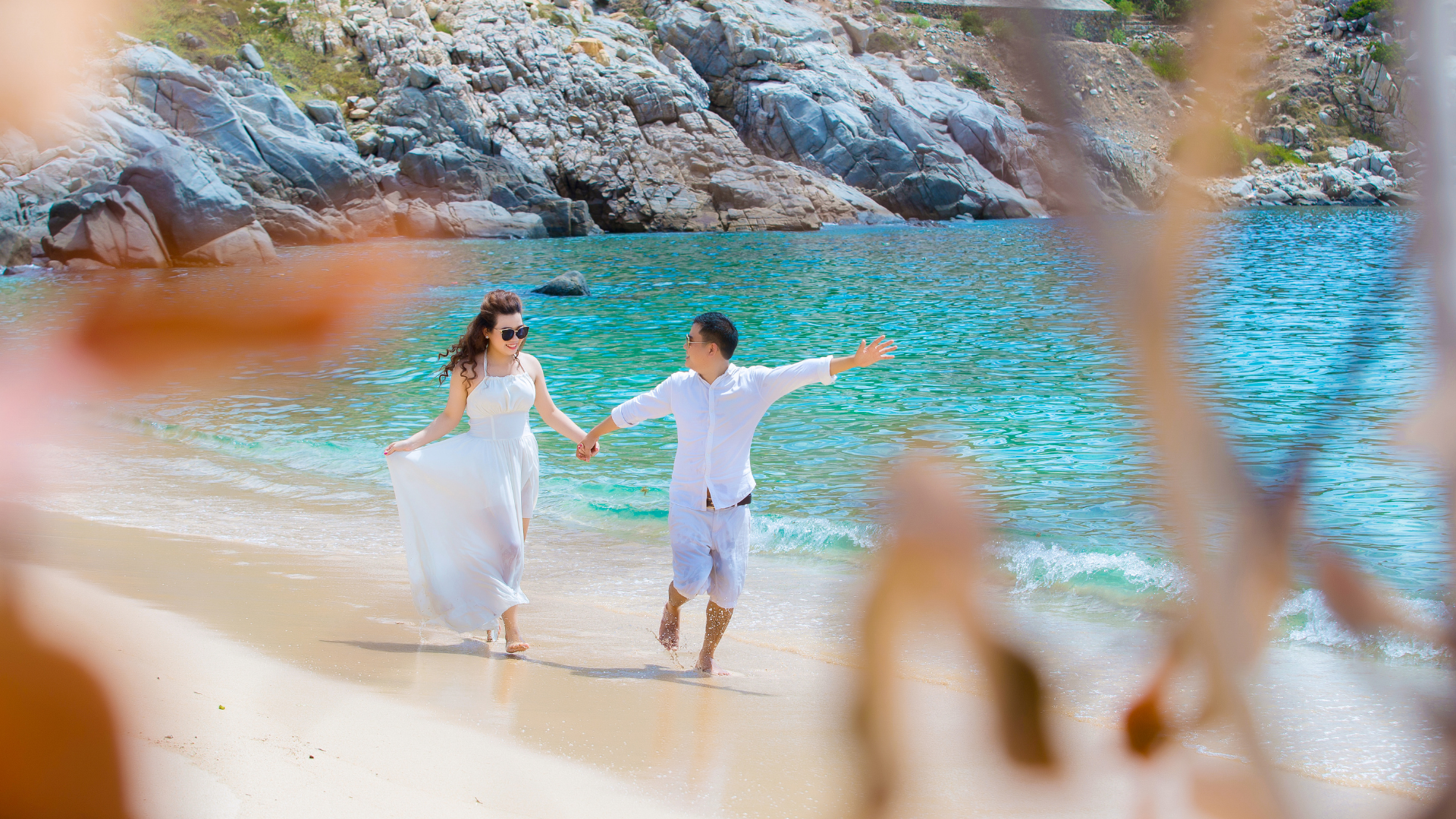 Couple holding hands on a sunny beach with turquoise water and rocky cliffs