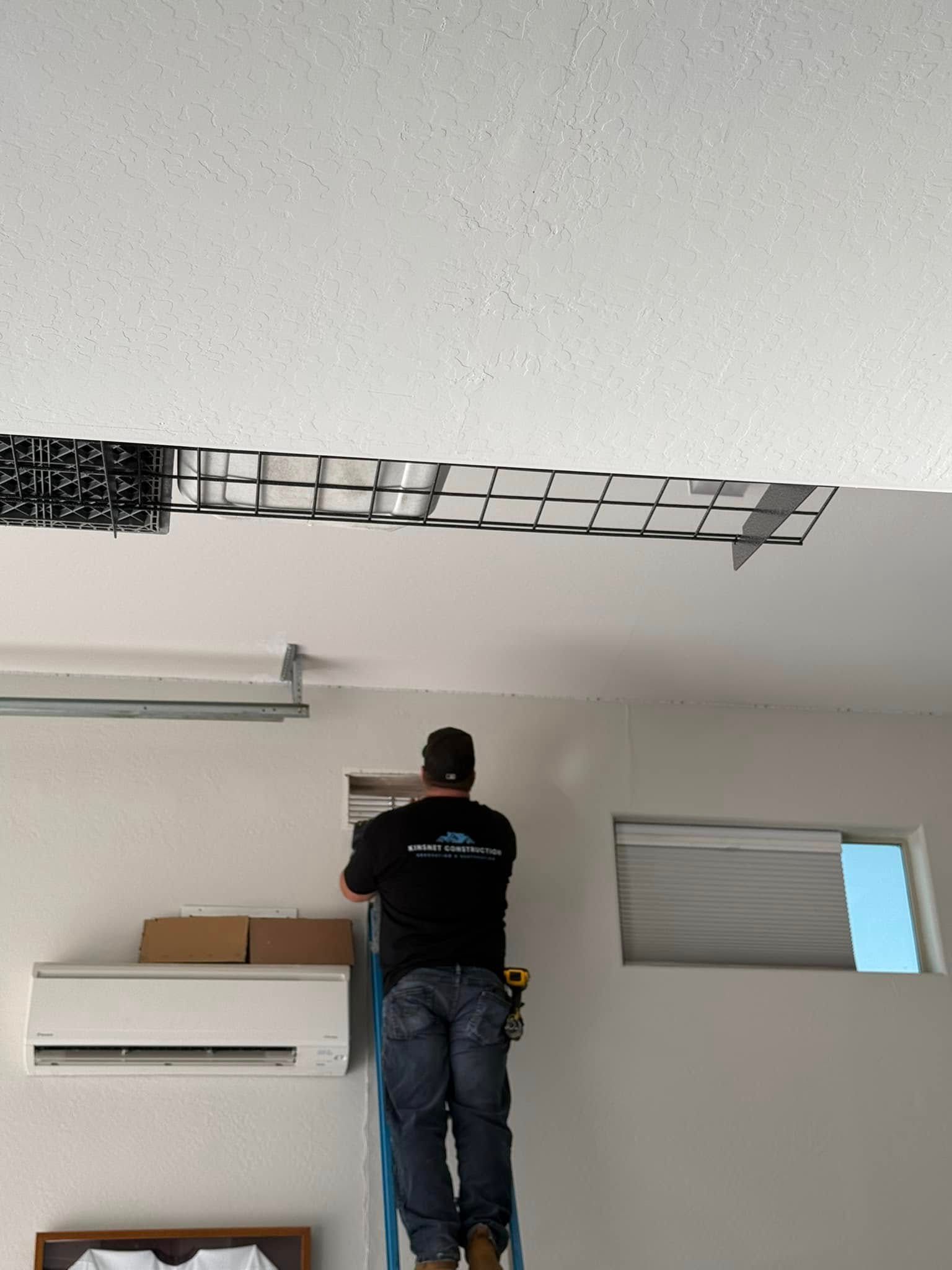 Man on ladder installs ductwork in a room with an air conditioner and window.