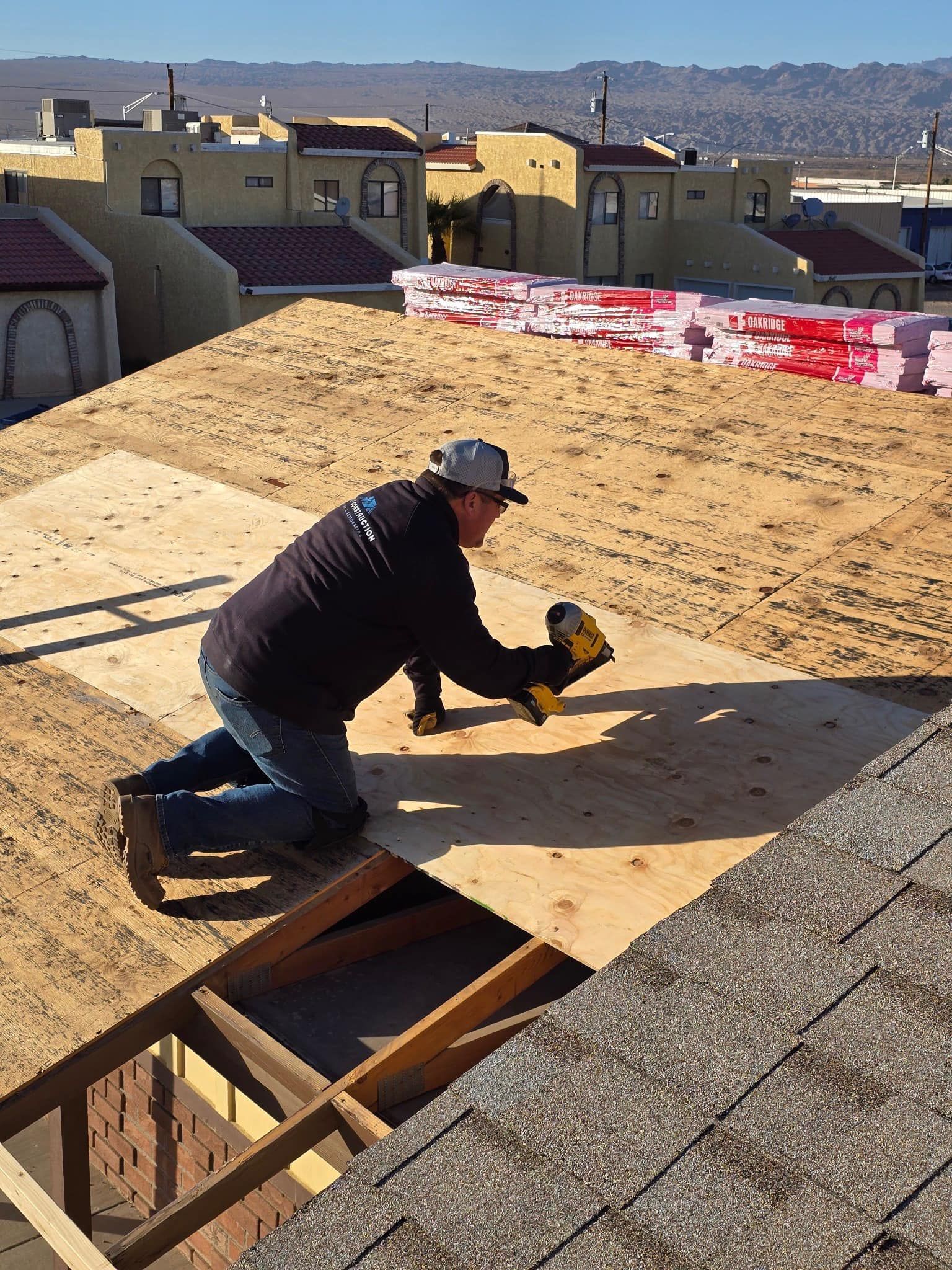 Roofer kneeling, nailing plywood to a roof. Houses and materials visible.