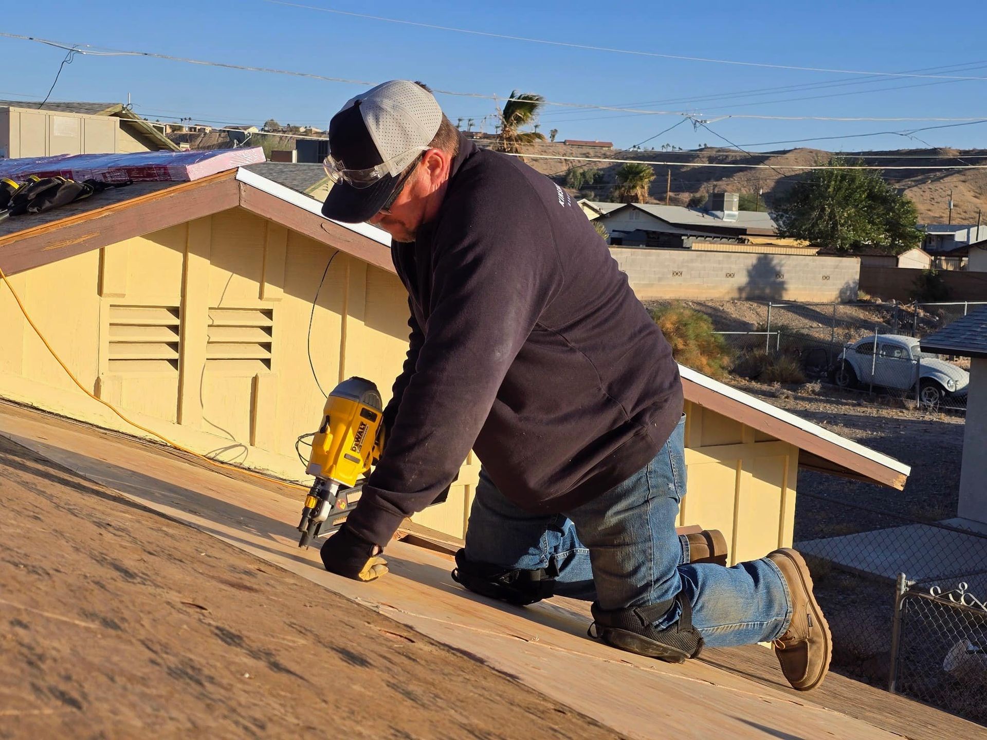 Roofer kneeling on a rooftop, using a nail gun. He's wearing a hat, knee pads, and jeans. Sunlight.
