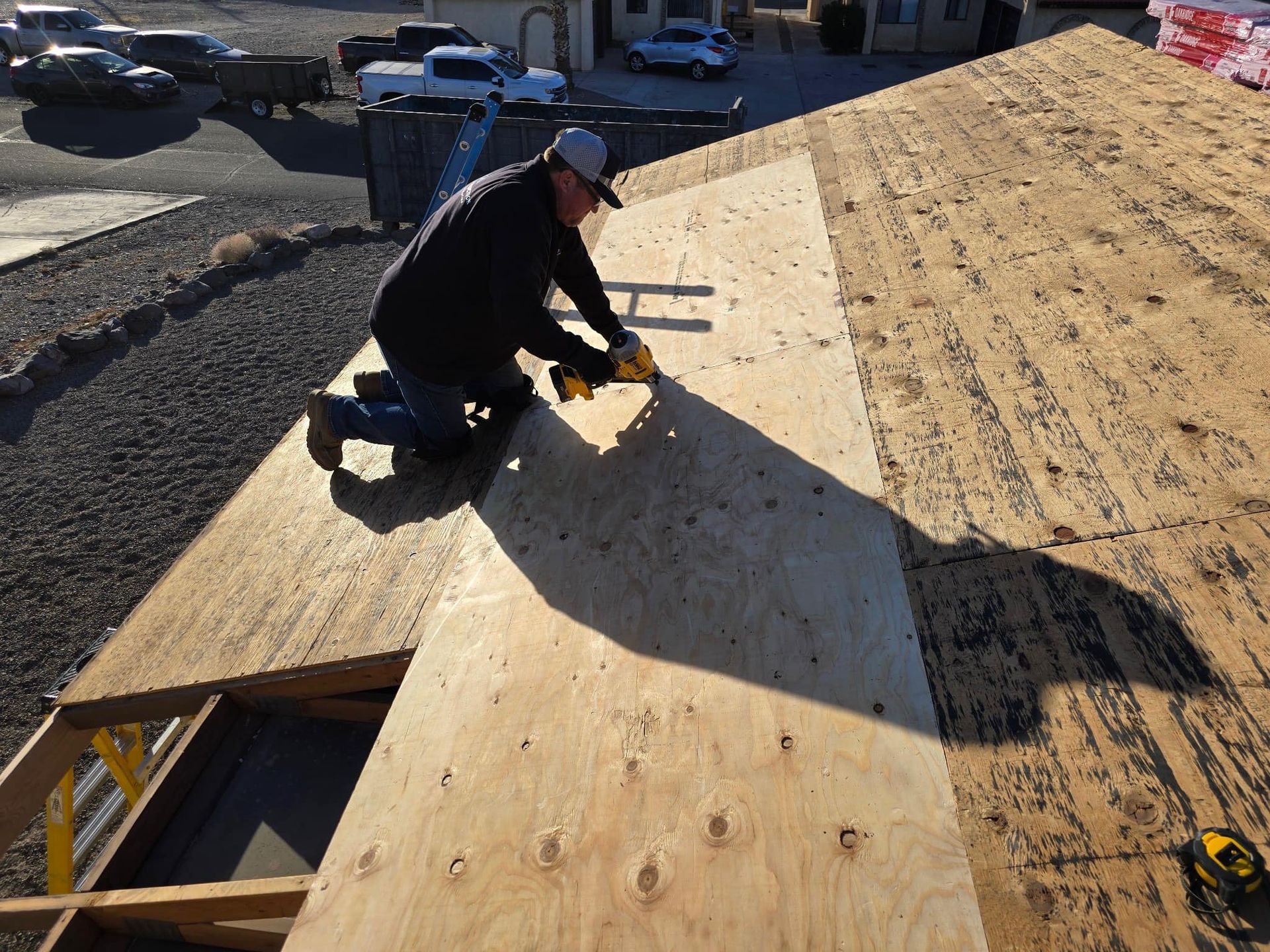 Person kneeling on a roof, using a drill to secure plywood. Sunlight casts long shadow.