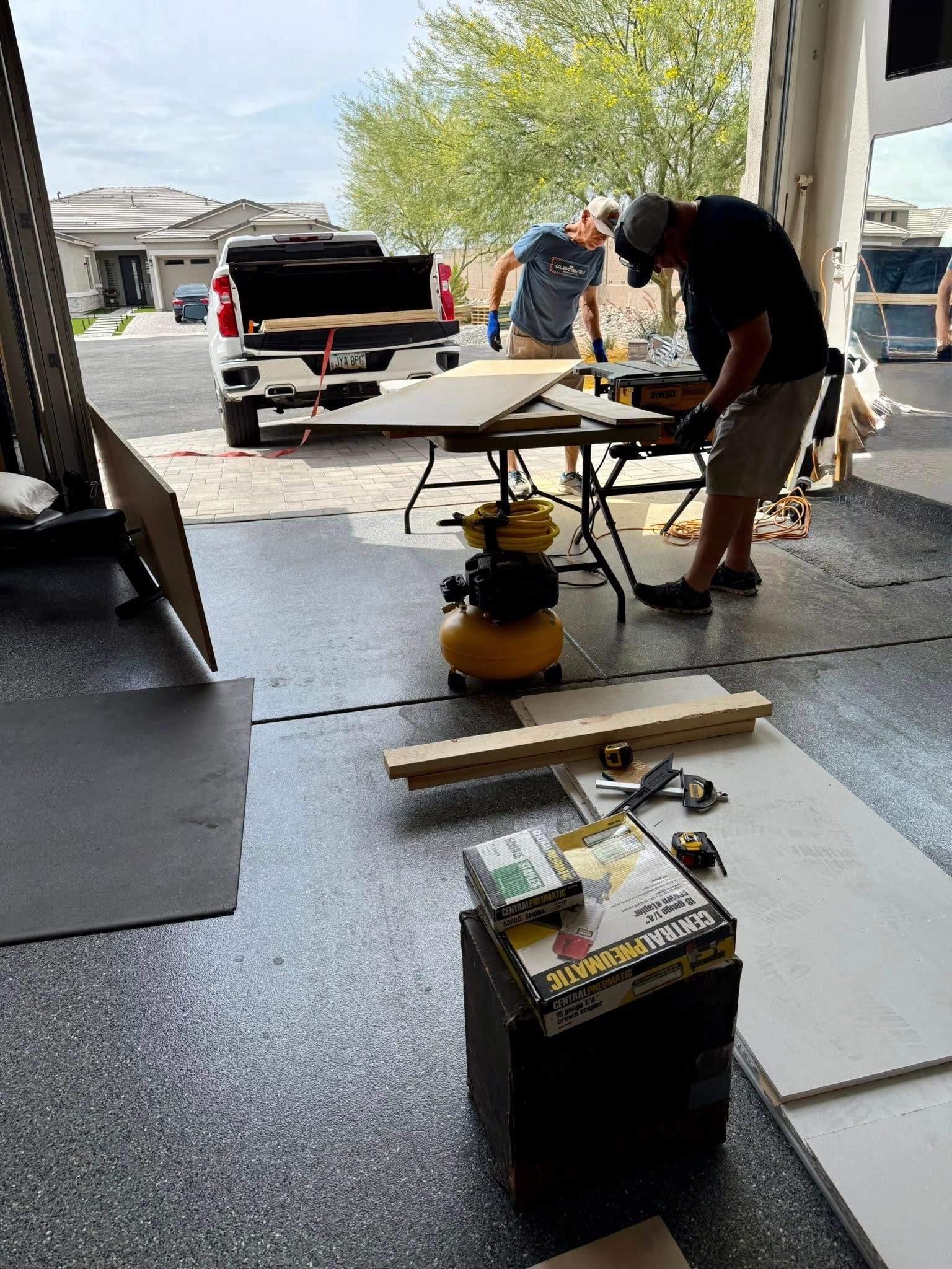 Two men cutting wood in a garage with a truck parked in the background. Tools and materials are scattered on the floor.