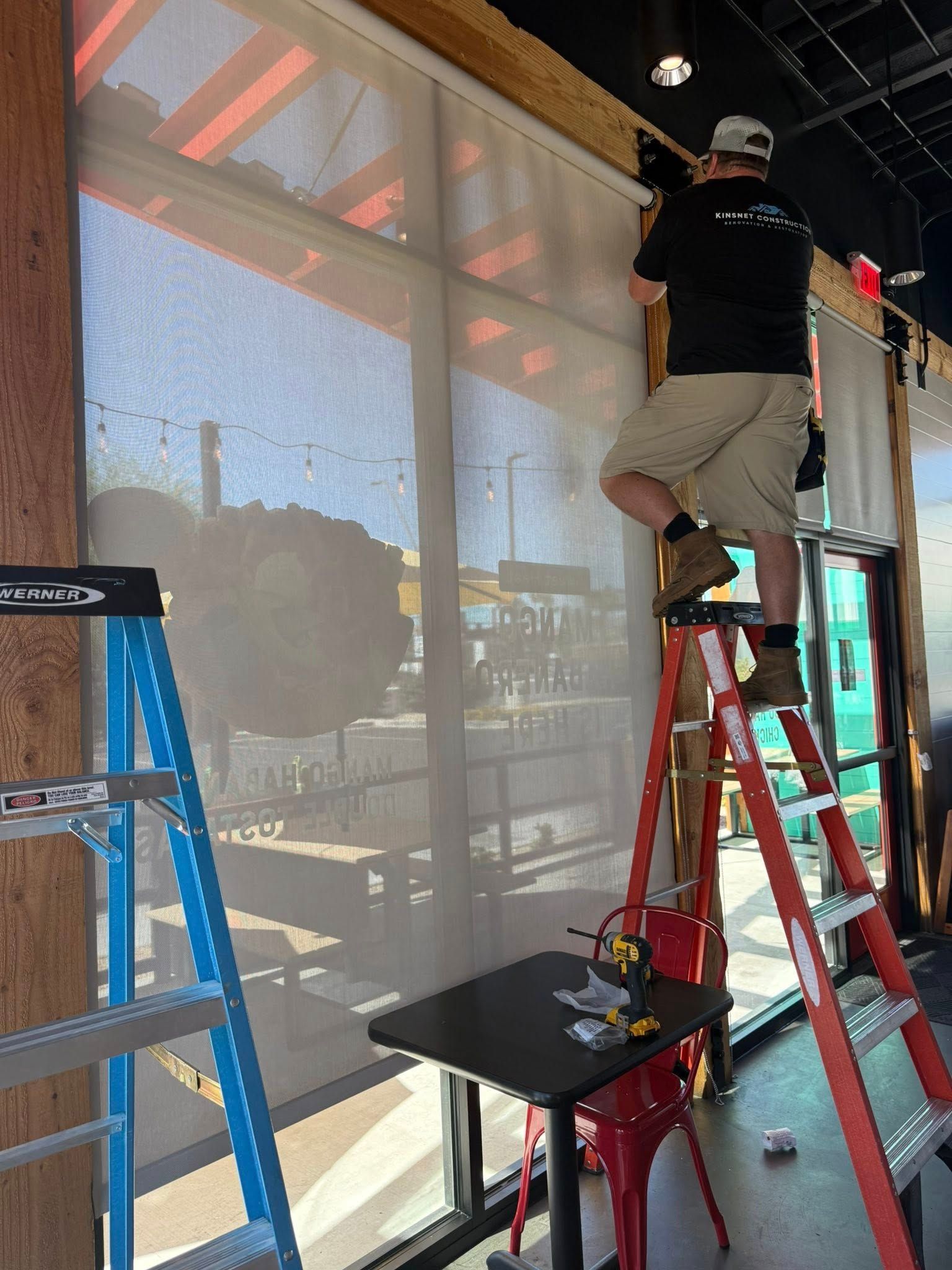 Man on a ladder installing a window shade in a restaurant. Includes table, ladder, tools, and window.