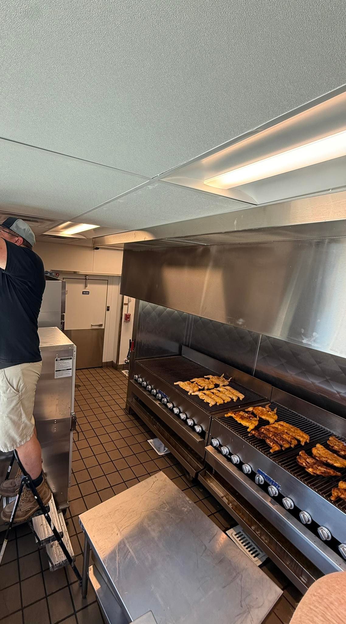 Kitchen with a grill cooking food, a person standing nearby, and a refrigerator.