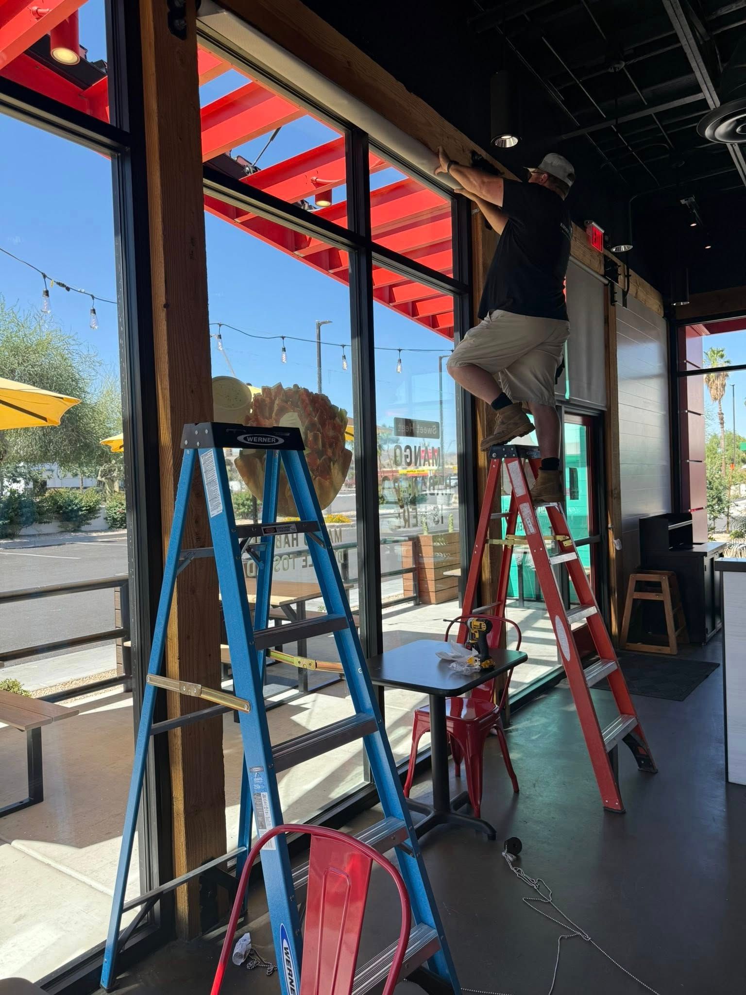 A person on a ladder working inside a restaurant with large windows.