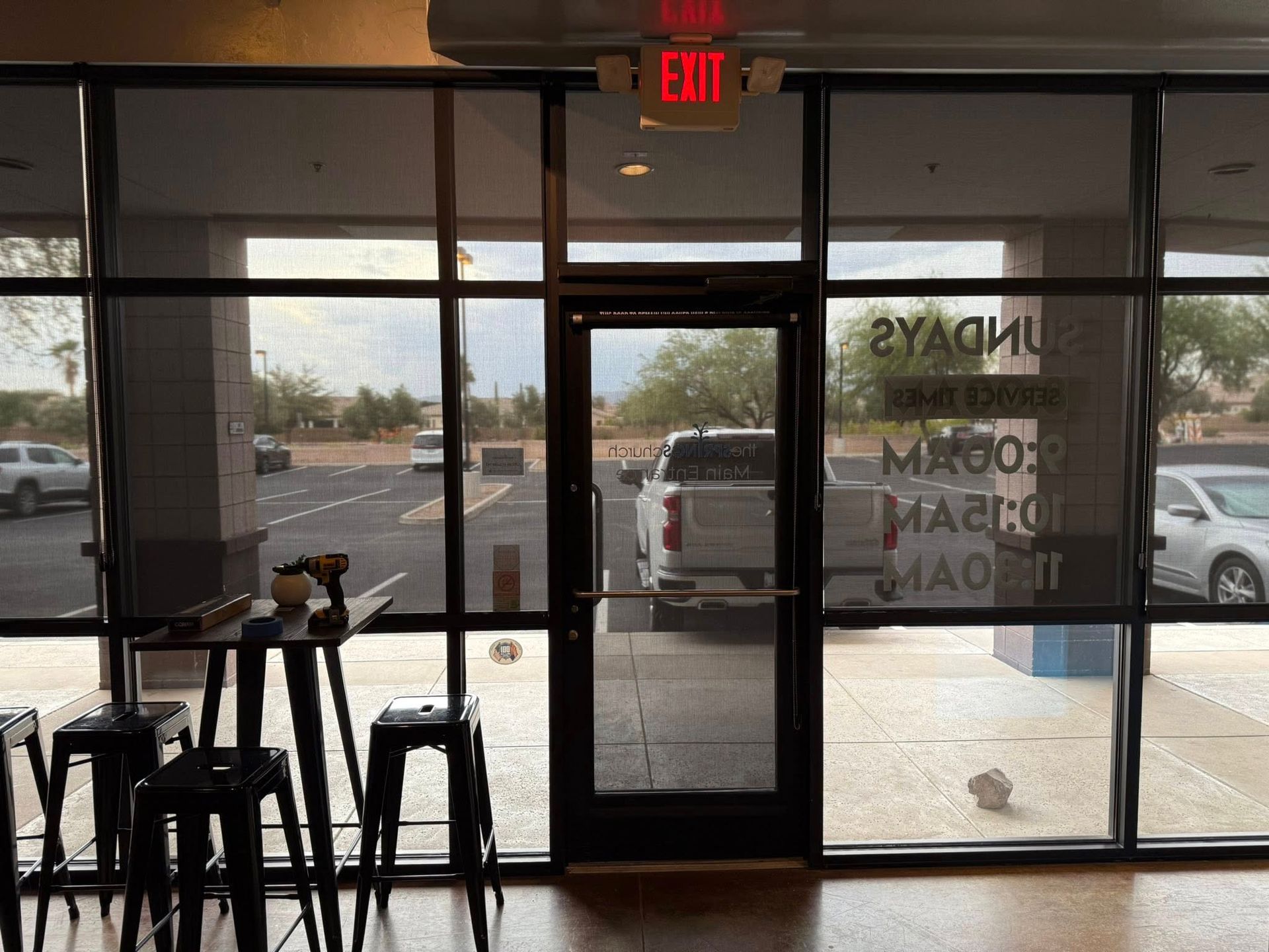 View of a business's front: door, windows, street, cars, tables, stools. Exit sign above door.