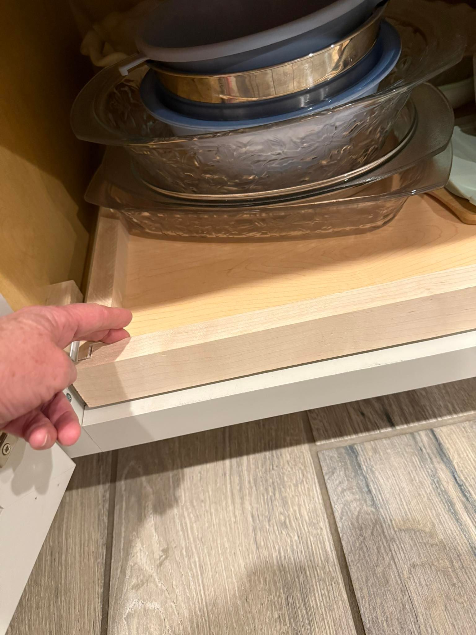 Person's hand pointing at pull-out shelf in cabinet holding dishes. Shelf is light wood, cabinet white.