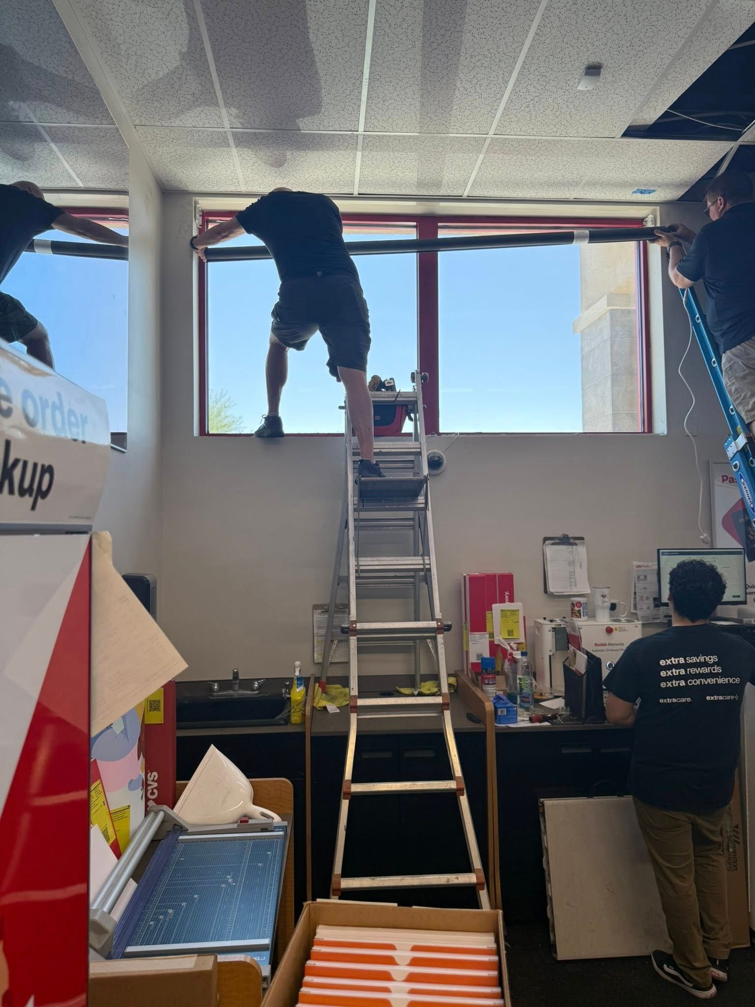 Three people installing something near a window inside a store. One on a ladder, two on ladders. Bright daylight outside.