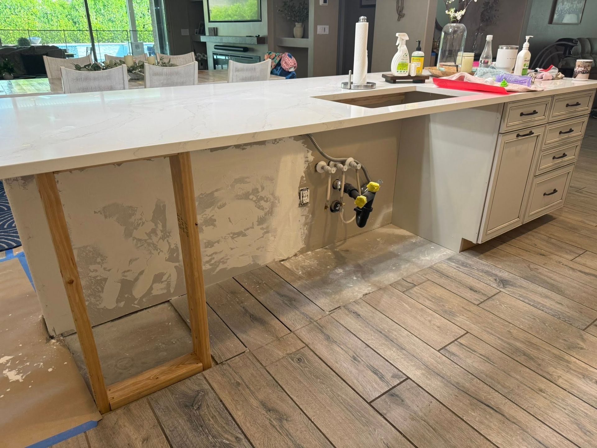 Kitchen island with exposed plumbing and unfinished drywall, wood-look tile floor.