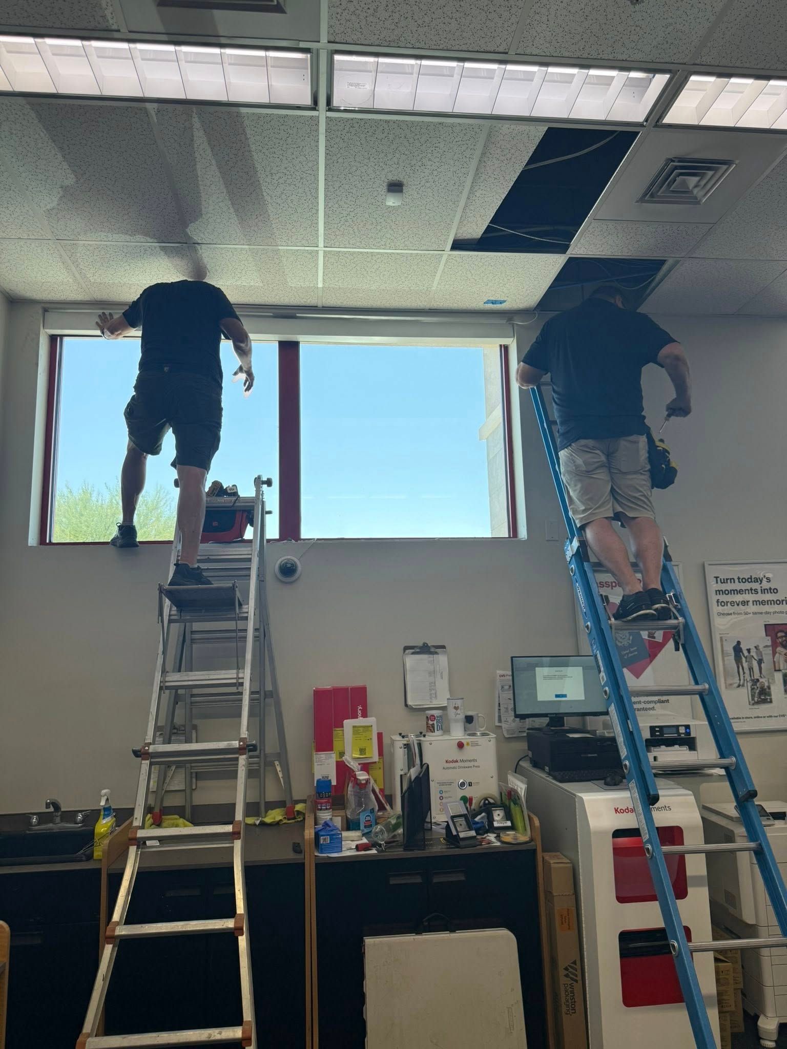 Two people on ladders working on a ceiling, near a window with a blue sky view.