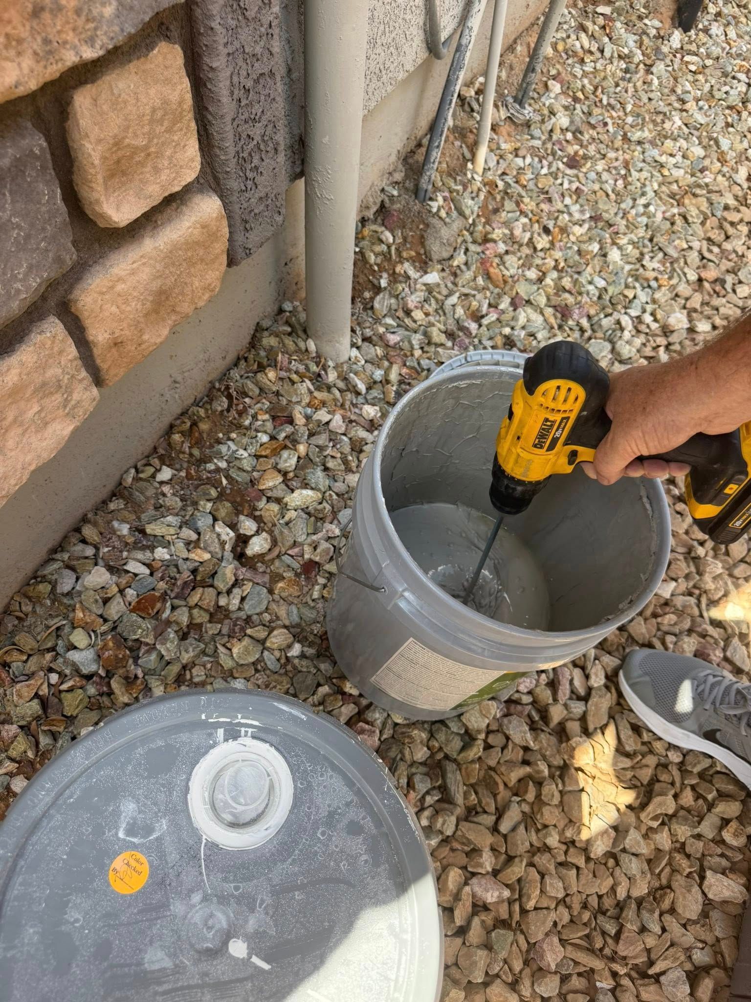 Person mixing a substance in a bucket with a power drill outside near a brick wall and gravel.
