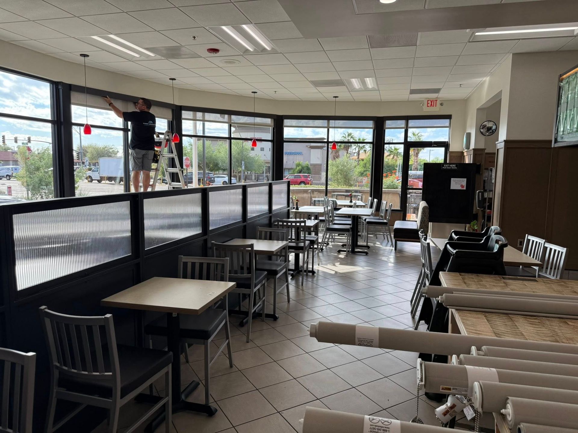 Interior of a restaurant. A person on a ladder installs fixtures. Tables and chairs are in place. Windows overlook a street.