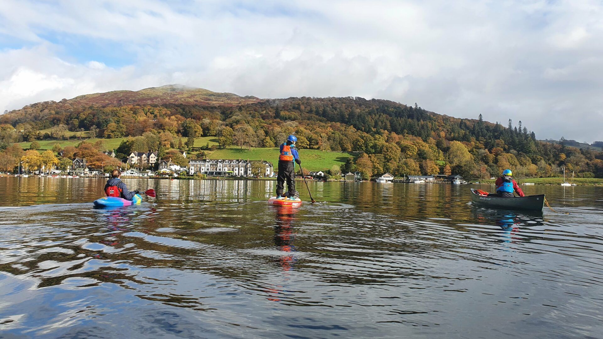 People paddleboarding and kayaking on a lake with a forested hill in the background, under a partly cloudy sky.