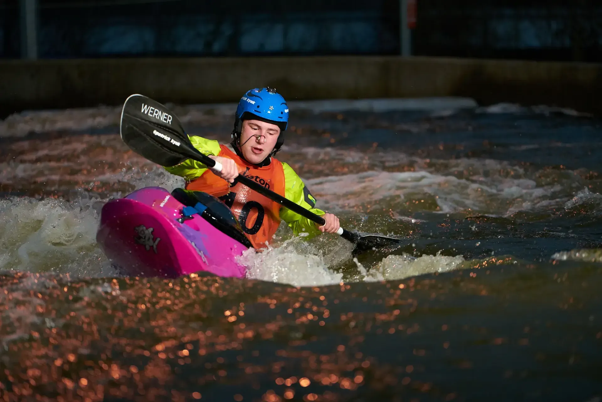 A kayaker wearing a blue helmet and colorful gear navigates whitewater rapids in a man-made channel, paddling forward with focused expression.